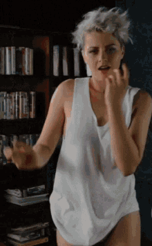 a woman wearing a white tank top is dancing in front of a bookshelf