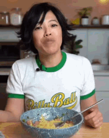 a woman wearing a billy ball t-shirt is eating a bowl of food .