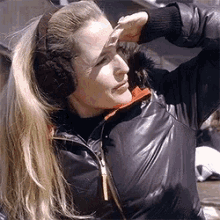 a woman wearing ear muffs and a leather jacket holds her hand to her forehead