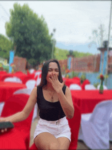 a woman in a black tank top and white shorts is sitting on a red chair covering her mouth with her hand