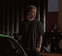 a man in a black shirt is standing next to a green car in a garage .