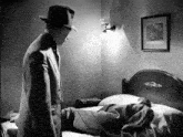 a black and white photo of a man in a suit and hat standing next to a bed