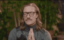 a man with long hair and glasses is praying with his eyes closed and his hands folded .