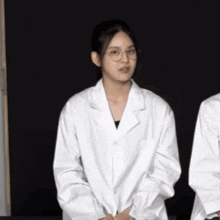 a woman wearing glasses and a white lab coat stands in front of a black background .