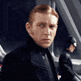 a man with red hair is wearing a black jacket and tie and looking at the camera .
