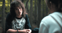 a man wearing a hellfire club shirt talks to another person