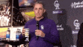 a man in a purple rockies jacket is holding a cup in front of a microphone