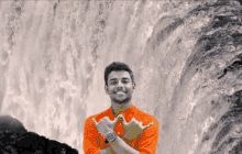 a young man in an orange shirt is standing in front of a waterfall