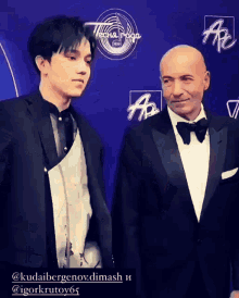 a man in a suit stands next to another man in a tuxedo