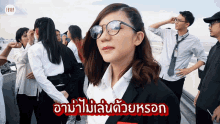 a woman wearing glasses and a suit stands in front of a crowd of people with foreign writing on the bottom
