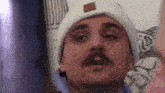 a man with a mustache wearing a white beanie and a blue shirt .