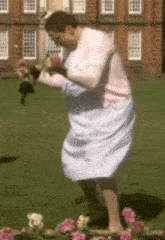 a woman in a pink sweater and white dress is standing in front of a brick building holding flowers .