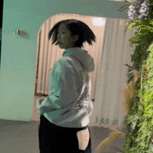a woman wearing a stussy hoodie is standing in front of a green wall of plants