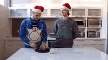 two men wearing santa hats and ugly christmas sweaters are standing in a kitchen preparing food .