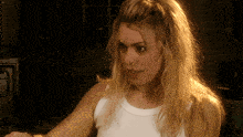 a blonde woman wearing a white tank top looks at something