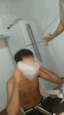 a shirtless man is sitting on the floor in front of a refrigerator while another man throws something at him .