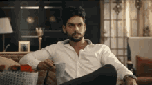 a man in a white shirt sits on a couch holding a cup