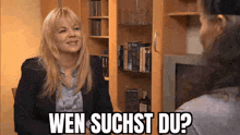 a woman is talking to another woman in a living room and the woman is smiling and asking , wen suchst du ?