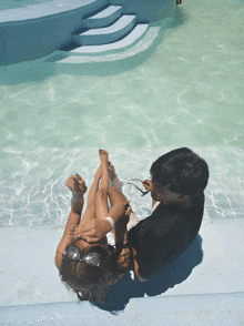 a man sits on the edge of a swimming pool with a woman laying on his lap