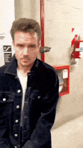 a man wearing a black denim jacket is standing in front of a red fire extinguisher .