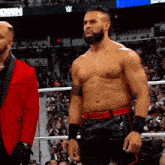 a shirtless wrestler in a red jacket stands next to another wrestler in a wrestling ring