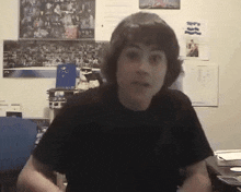 a young man in a black shirt is sitting in front of a wall with posters on it