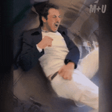 a man in a suit and white pants is laying on a bed with m + u written on the bottom right corner