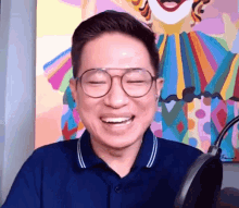 a man wearing glasses and a blue shirt is laughing in front of a clown painting .