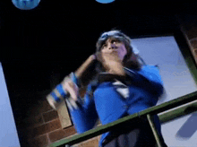 a woman in a blue jacket and goggles is standing on a balcony holding a hammer .