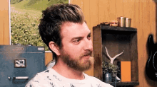 a man with a beard is sitting in a room in front of a shelf with a picture on it .