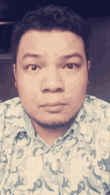 a man wearing a shirt with a floral pattern looks at the camera with a serious look on his face