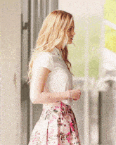a woman wearing a white shirt and a pink floral skirt stands in front of a window