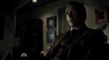 a man in a suit and tie is sitting in a dark room with a nbc logo in the corner .