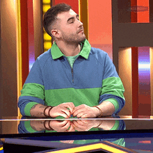 a man in a blue and green sweater is sitting at a table with his hands on the table and the word pass behind him