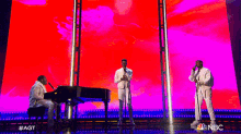 three men singing and playing a piano on a stage with a nbc logo in the background