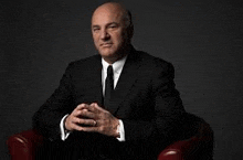 a man in a black suit and tie is sitting in a red chair with his hands folded .