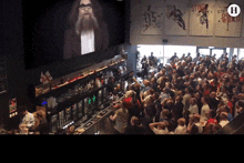 a large crowd of people watching a man on a large screen with the letter h in the middle
