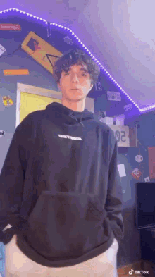 a young man in a black hoodie is standing in a room with his hands in his pockets .
