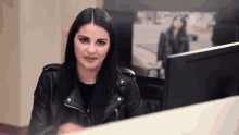 a woman in a black leather jacket is sitting at a desk in front of a computer .