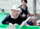 two men are sitting on a green boat and one of them is wearing a bucket hat