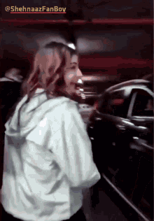 a woman in a white jacket is standing in front of a car with a caption that says shehnaazfanboy on it