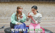 two girls are sitting on a bench eating food in front of a brick wall .