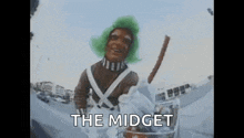 a man in a green wig is riding a bicycle with a stick in his hand and the words `` the midget '' .
