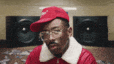 a man wearing a red hat and glasses looks at the camera in front of a speaker