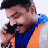 a man with a mustache is wearing an orange vest and talking on a cell phone .
