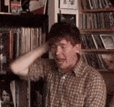 a man in a plaid shirt is sitting in front of a bookshelf with his hand on his head .