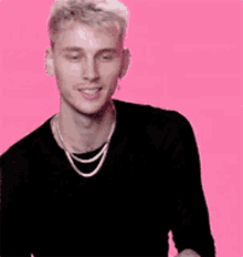 a young man wearing a black shirt and a silver chain around his neck is smiling on a pink background .