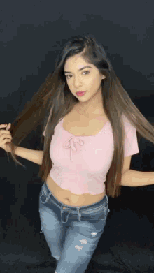 a woman in a pink crop top and blue jeans holds her hair