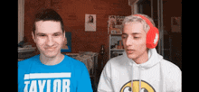 a man wearing a blue shirt that says taylor is standing next to another man wearing red headphones .