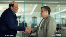two men shaking hands in front of a glass wall with the nbc logo in the corner